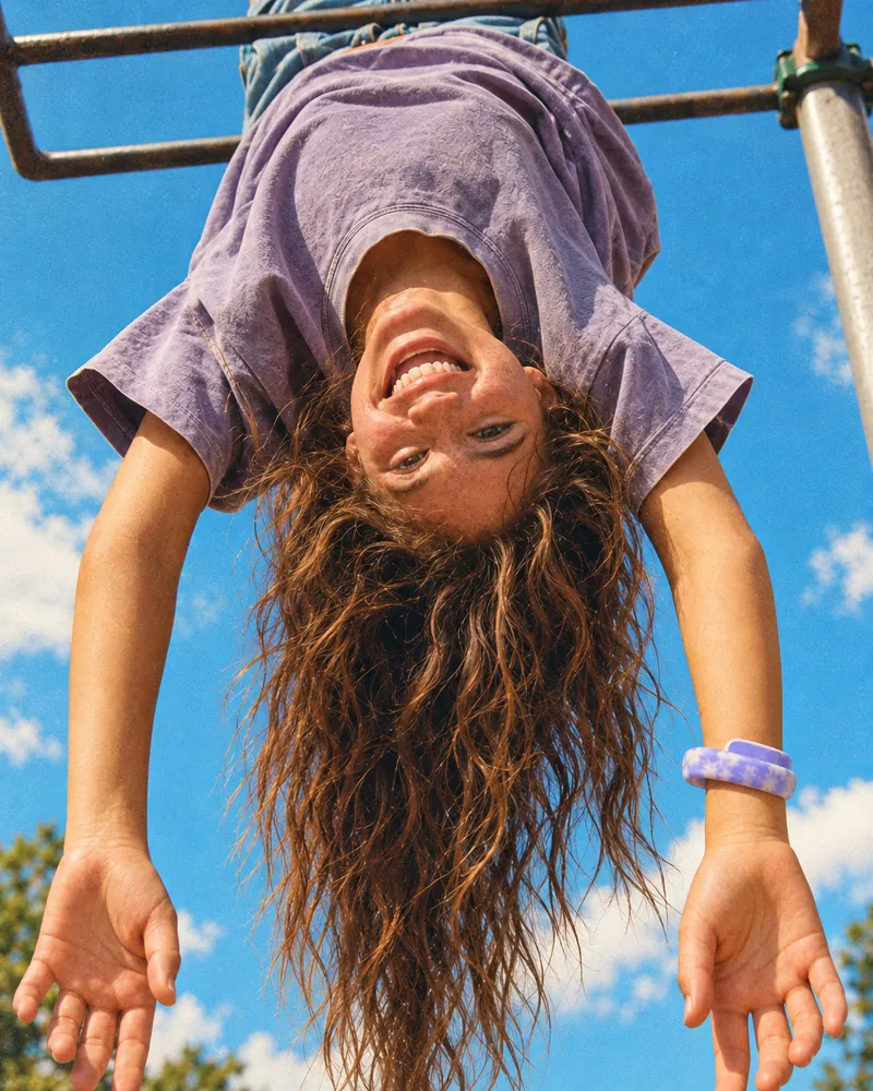 Lola hanging upside down from the monkey bars, laughing, a lilac Sky Loop on her wrist