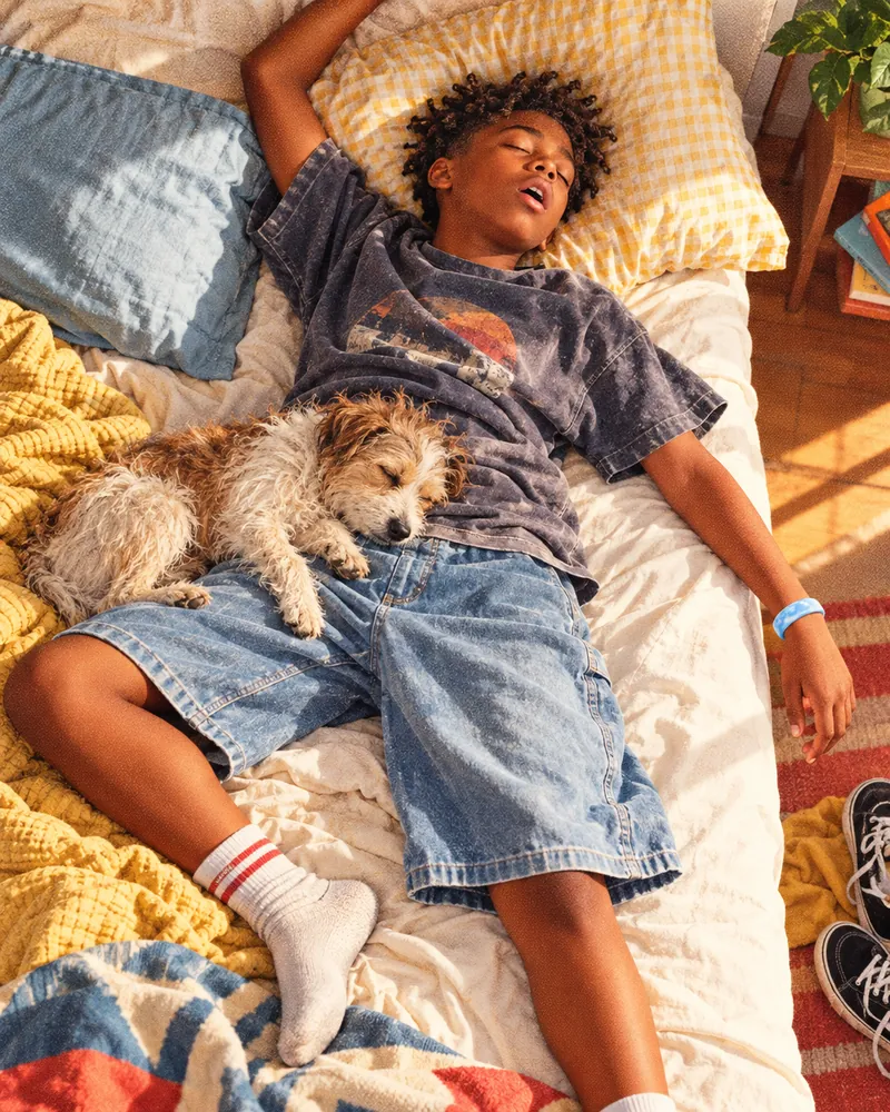 A boy fast asleep across a messy bed, his dog asleep on his legs, a glacier Sky Loop on the arm flung off the mattress