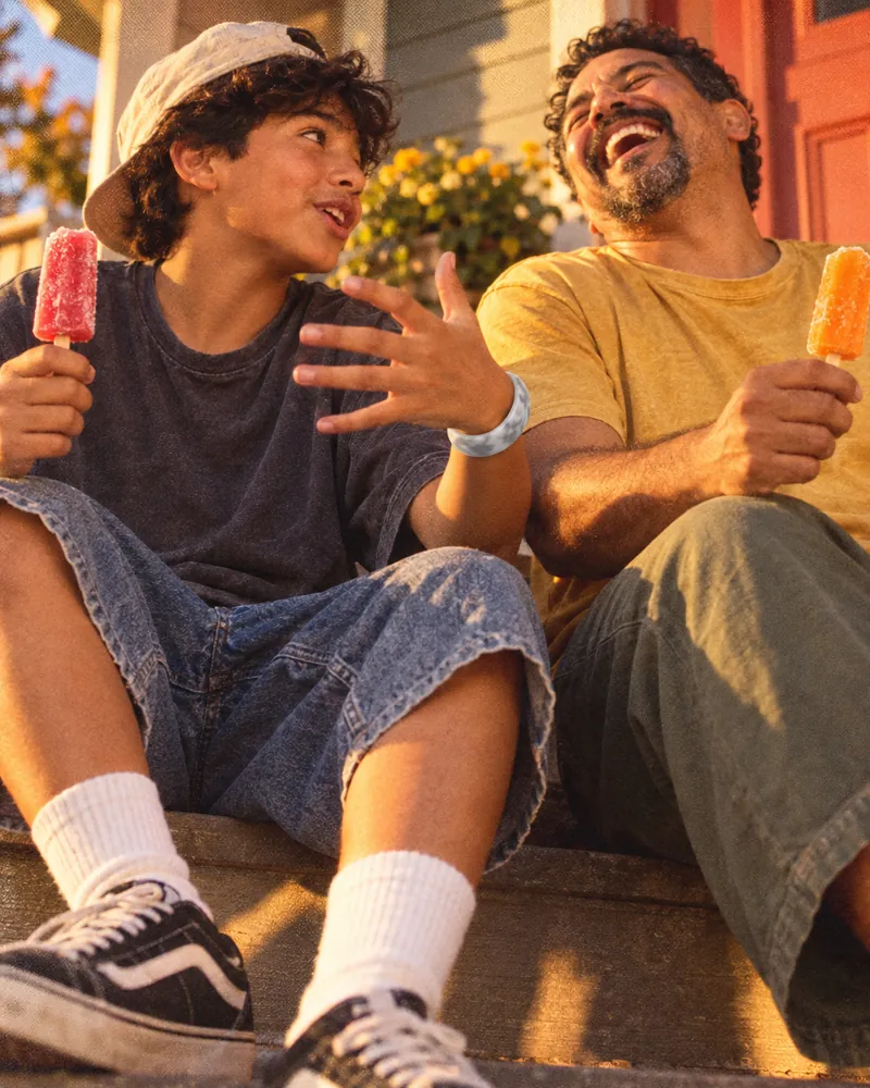 A boy telling his dad a story on the front steps, both holding ice lollies, the dad laughing, a smoke Sky Loop on the boy’s wrist